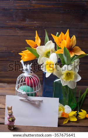 Colorful easter eggs in bird cage, fresh tulips and narcissus flowers  in vase, empty tag  on aged dark wooden background. Easter background. Place for text.
