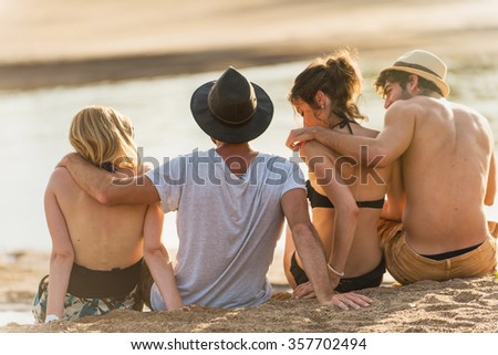 Back view of two young couple in their 30's at the beach. They are sitting in the sand arm in arm, enjoying the last lights of the day and looking at the ocean. They are wearing swimsuits and hats.