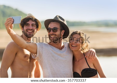 Three friends in their 30's taking selfies at the beach. A woman wearing a bikini and two men with hats and sunglasses are having fun with a smartphone. They are looking at the screen for the picture.