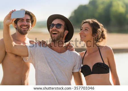 Three friends taking selfies at the beach. A woman wearing a bikini and two men with hats and sunglasses are having fun with a smartphone. They are looking at the screen for the picture.