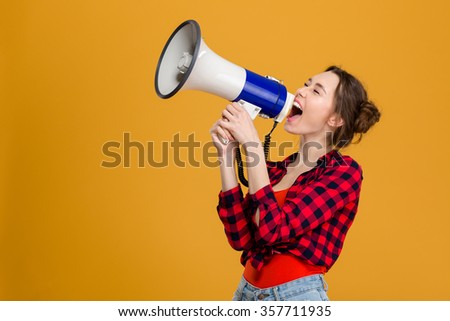Funny excited young woman in checkered shirt shouting in megaphone over yellow background Royalty-Free Stock Photo #357711935