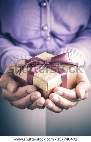 Close up of hands holding a small gift wrapped with purple ribbon. Small gift in the hands of a woman indoor. Shallow depth of field with focus on the little box.