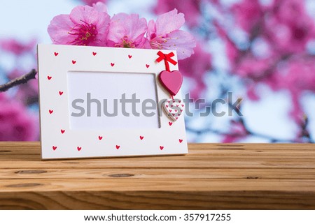 White photo frame with red heart on wooden table over pink cherry blossoms flowers background