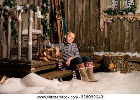 Portrait of cute little boy in boots on the background of the porch and stacked firewood