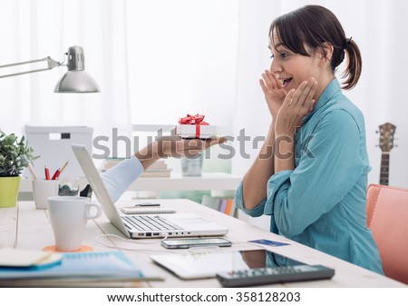 Cheerful young woman connecting to internet and receiving a present from a website, a male hand is coming out from the computer screen and giving her a gift box Royalty-Free Stock Photo #358128203