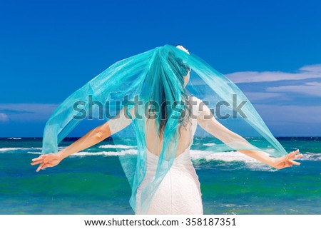 Beautiful brunette bride in white wedding dress with turquoise veil stand on shore sea. Tropical sea in the background. Summer vacation concept.