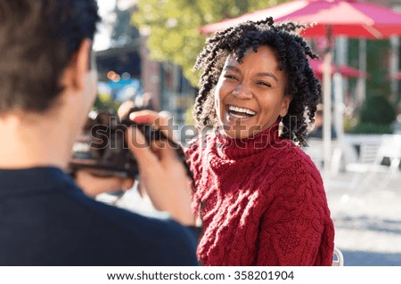 Young beautiful african woman posing for a photo. Boyfriend clicking a picture with camera of an smiling girlfriend. Happy woman having photos clicked by a photographer outside. Man taking a picture.