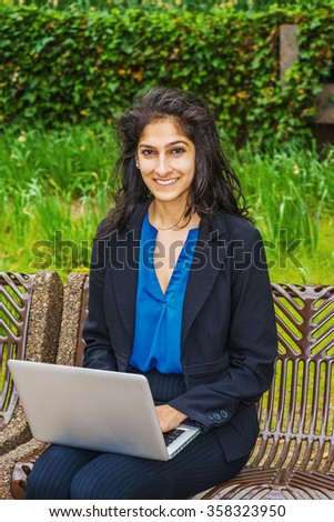 Happy East Indian American college student studying in New York, wearing black blazer, blue shirt, striped pants, sitting on chair on campus, reading, working on laptop computer, smiling, enjoying.
