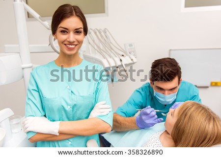 Portrait of beautiful assistant, crossed hands, against dentist treats teeth little girl Royalty-Free Stock Photo #358326890