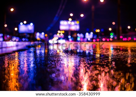 Rainy night in the big city, the light from the headlamps of vehicles approaching on the highway. View from the level of asphalt, image in the soft orange-purple toning