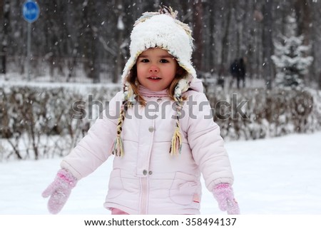Happy toddler girl in warm coat and knitted hat tossing up snow and having a fun in the winter forest, outdoor portrait