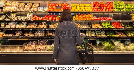 Woman standing in front of a row of produce in a grocery store.  Royalty-Free Stock Photo #358499339