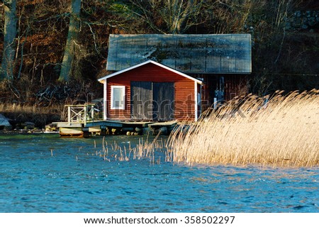 An old red, wooden boathouse with a small jetty in front. Reed and water in foreground and forest in background. Wind is bending the reed. Late December in Jarnavik bay outside Ronneby, Sweden. Royalty-Free Stock Photo #358502297