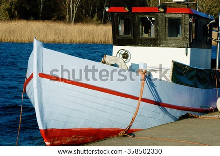 A small red and white fishing boat moored at dockside with sea and reed in the background. The cabin is black with sunlight reflections. Late December in Jarnavik bay outside Ronneby, Sweden. Royalty-Free Stock Photo #358502330