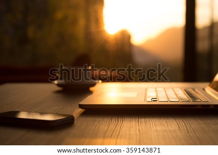 Nomad Work Concept Image Computer Coffee Mug and Telephone on black wood Table and Evening Sunlight shining throw large windows focus on Edge of Laptop Royalty-Free Stock Photo #359143871