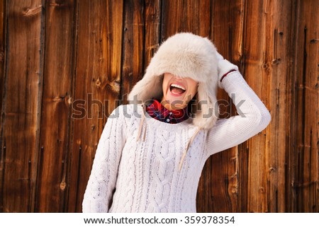 Winter in the country style. Smiling young woman in white knitted sweater pulled furry hat over her eyes in the front of rustic wood wall