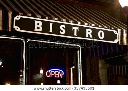 Striped awning with sign reading Bistro over a window with a string of lights around it and a neon open sign