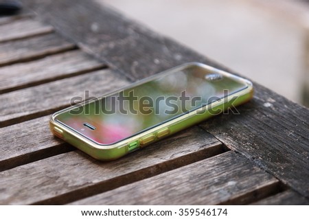 old green smartphone in a transparent silicone case laying on an old wood table with rustic weathered texture outdoor reflecting colorful natural garden environment vivid bokeh on the touch surface