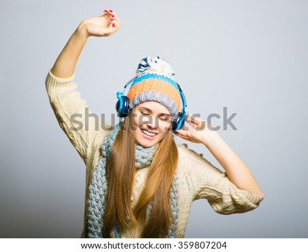 beautiful girl happy listening to music with big headphones with a phone or player, knit cap, winter concept, photo studio, portrait of a woman isolated on gray background