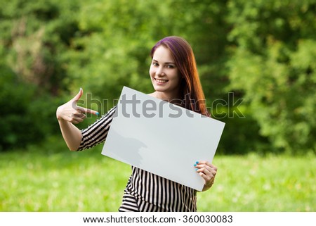 Beautiful young woman in a striped t-shirt with colored hair and nails holding a blank white board and pointing and it, outdoors on natural background