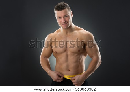 Muscular man measuring waistline with measure tape against black background