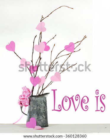 Paper cut out heart shapes in two shades of pink on bare twigs in a rustic tin pitcher sitting on a white washed wooden table on a bright background. Vertical composition with love is text message 