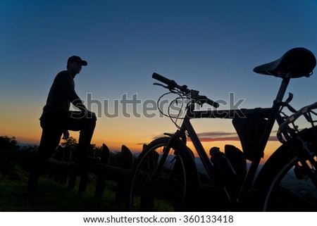 Silhouette of Half Profile cyclist on top of mountain with sunset in background. Male next to mountain bike bicycle wearing a baseball hat and resting one foot on a rail and leaning on his knee.