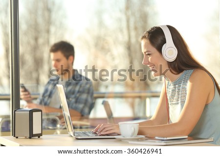 Side view of a beautiful female working on line with headphones typing in a laptop in a restaurant indoor with a window in the background and people outdoors