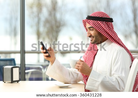Side view of an arab man texting in a smart phone in a coffee shop with a window with a sunny day in the background