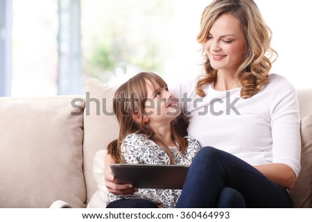 Portrait of a beautiful happy mother and her cute daughter sitting in a beige sofa together and using digital tablet. 