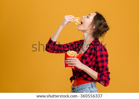 Profile of beautiful happy woman in plaid shirt tasting french fries over yellow background Royalty-Free Stock Photo #360556337