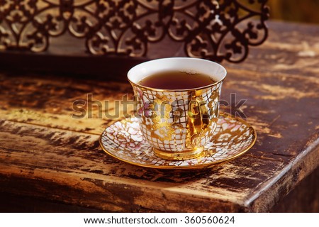 Closeup image of vintage porcelain tea cup on saucer with golden floral pattern at wooden desk background.