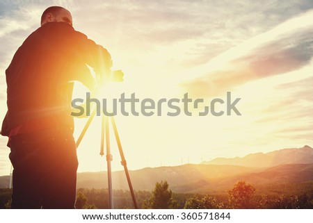 Silhouette of a man geo-desist using theodolite for determines the required coordinates to construct a new building structure, male surveyor measures the distance while standing against sunset