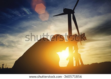 Silhouette of a man geo-desist using theodolite for measures distance for bridge construction while standing against windmills and sunset, male surveyor determines the required coordinates