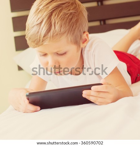 Child lies on bed and plays games on tablet. Inside. Toned image with sun light effect. Theme of modern technologies