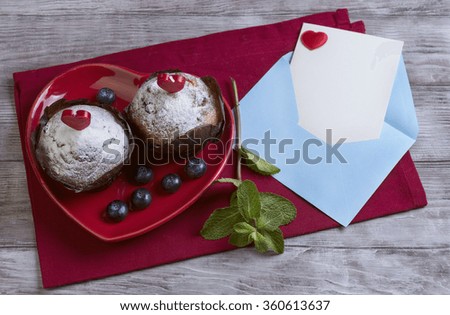 On wooden table in a bright red cloth with plate in the form of heart with two berry muffins with candles, sugar, blueberry, mint, envelope and sheet of paper, card Happy Valentine's Day, top view