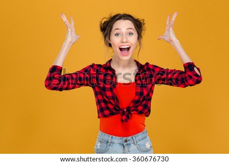 Happy successful excited beautiful young woman in plaid shirt shouting with raised hands over yellow background