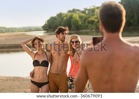 Four friends in their 30's are standing close to each other in the sand at the beach, wearing bikinis, shorts, sundresses and hats. One of them is taking a shot of the three others with a smartphone.