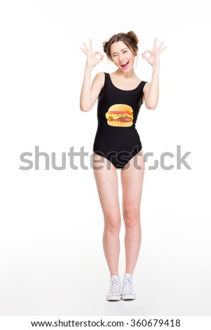 Smiling charming young female in black swimsut with hamburger print and white sneakers standing and showing ok sign over white background
