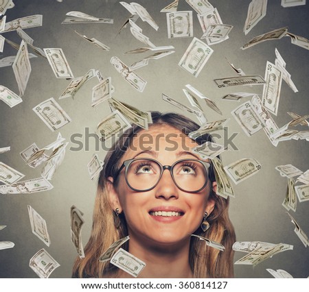 Portrait happy successful young woman in glasses looking up under a money rain dollar bills banknotes falling down isolated on gray wall background with copy space  