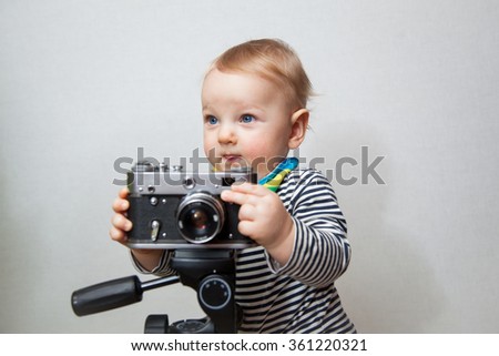 Little beautiful baby boy with smiley face holding vintage film photocamera on a light gray wall background