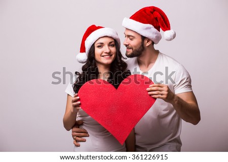 Romantic Young Happy Couple with santa hat hugging and big red heart in hands. He looks at she. isolated on grey background