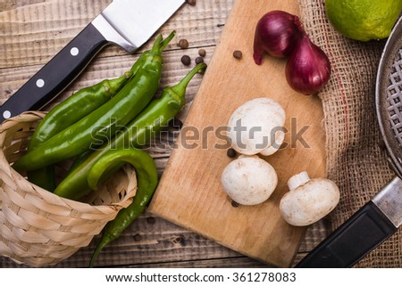 Fresh white mushrooms sweet onions on cutting board overturned pottle with green chili peppers lime with pan on burlap and knife on table, horizontal picture