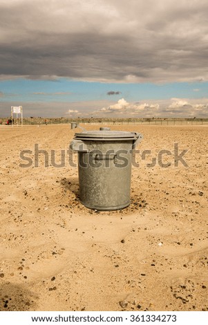 Garbage can in the sand at the beach. Cloudy sky.