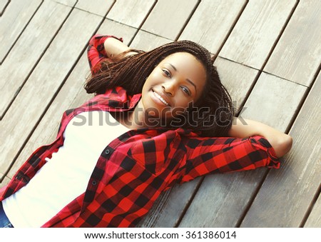 Beautiful smiling young african woman relaxed on wooden floor with hands behind head, wearing a red checkered shirt, top view