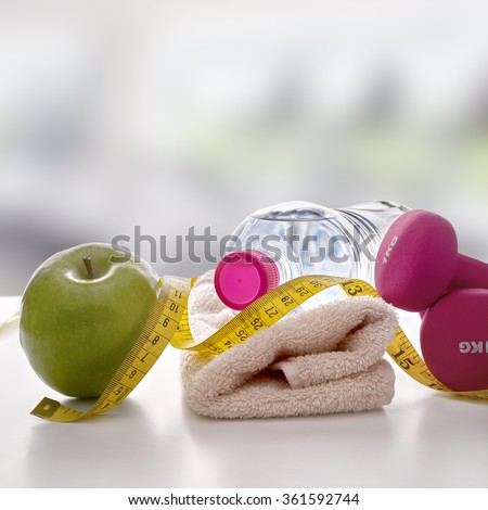 Pink dumbbells with apple, mineral water bottle, towel and tape measure on white table and gym background. Concept health, diet and sports. Square composition. Front view
