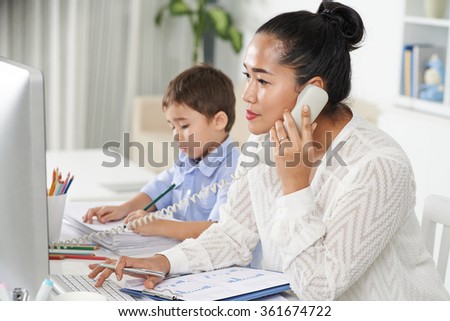 Concentrated female entrepreneur calling on the phone when her son drawing on documents
