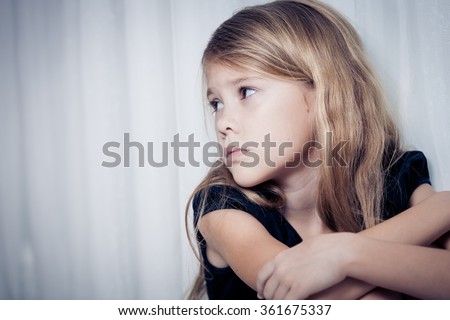 Portrait of sad little girl sitting near the window at home at the day time