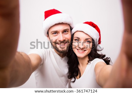 christmas, holidays, technology and people concept - happy couple in santa hats taking selfie picture from hands on white background. They look at camera.
