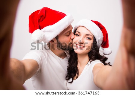 christmas, holidays, technology and people concept - happy couple in santa hats taking selfie picture from hand on white background. He kiss she.